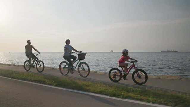 Happy family of three enjoying a beautiful morning by the sea, a little girl riding a bike with her mom and dad, handheld shot.