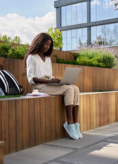 Happy black young woman model, pretty curly African female university student elearning using laptop computer studying outdoors in park on sunny day, remote learning online, vertical.