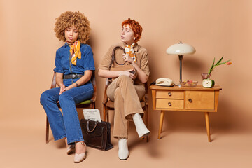 Two young women wait in queue near cabinet sit on chairs talk to each other dressed in elegant clothes isolated over brown background. Retro vintage style. Horizontal shot. Back to 80s concept