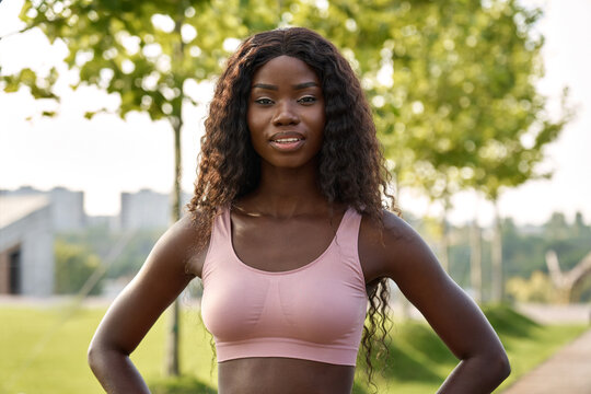 Young adult confident beautiful happy fit sporty slim active black African ethnic woman model wearing sportswear top standing in city park outdoors looking at camera. Close up portrait.