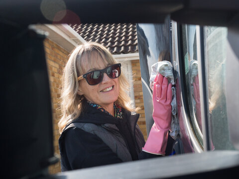 An attractive lady smiling at camera is framed through wing mirror brackets using a cloth to wipe the side of her motorhome recreational vehicle.She wears pink rubber gloves and sunglasses