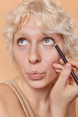 Headshot of blonde young woman applies eyeliner on eyelid with pencil focused above keep lips rounded undergoes beauty procedures wants to look beautiful poses against brown studio background.