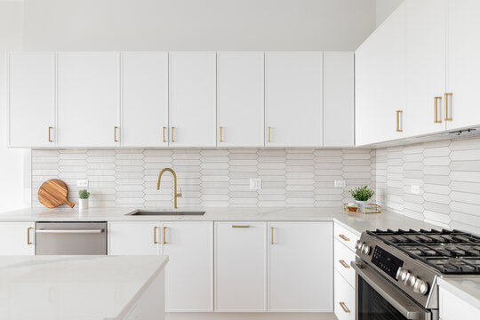 A Beautiful Kitchen Detail With White Cabinets, A Gold Faucet, White Marble Countertops, And A Brown Picket Ceramic Tile Backsplash.