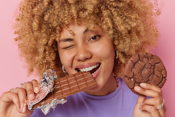 Close up shot of beautiful woman with curly hair bites bar of chocolate holds cookie winks eye has...