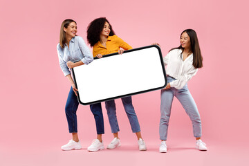 Excited diverse female friends holding huge smartphone with empty screen on pink studio background, mockup