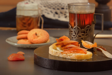 Piece of rustic bread with pear jam, a glass of hot tea in an antique silver cup holder and a jar of jam on a gray wooden table. Minimalism. Village breakfast