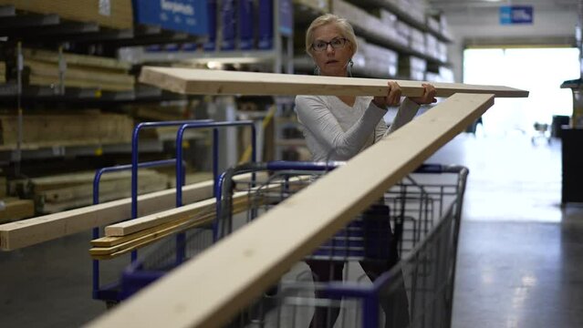 Mature woman putting construction wood in shopping cart in the lumber section of a big box hardware store.
