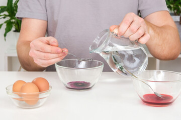 A man pours spoonful water into glass cup with dye. Dyeing eggs with coloring in Easter egg