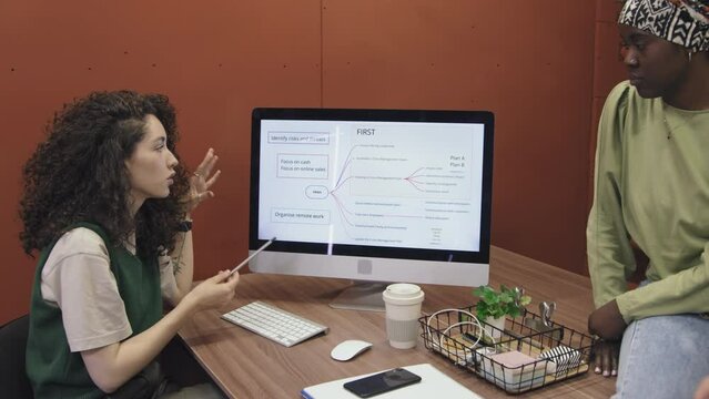 Medium shot of energetic young Caucasian female startup manager sitting at desk in shared office facility, demonstrating her business growth strategy to colleagues and talking persuasively