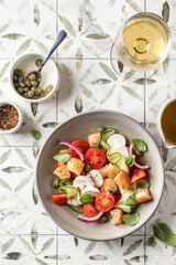 Traditional italian tomato salad panzanella with mozzarella, capers, red onion, croutons, cucumbers and basil with fork and wine glass. Summer salad on printed tile background top view