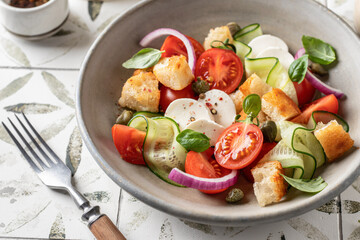 Traditional italian tomato salad panzanella with mozzarella, capers, red onion, croutons, cucumbers and basil. Summer salad on printed tile background, close up