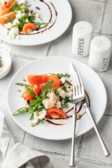 Healthy vegetable salad with fresh arugula, tomato, feta cheese on white plate with cutlery, gray tile background. Diet menu with text space