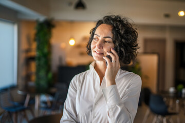 One woman mature caucasian female businesswoman entrepreneur stand at work or home use mobile phone making a call talk real people copy space wear white shirt curly hair happy smile