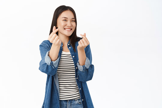 Cute young woman, 20 years old, asian girl shows heart finger sign, I love you gestures, stands over white background