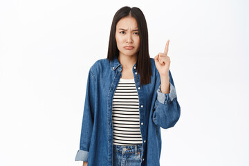 Upset sulking asian girl points finger up, grimaces from smth unfair, complains, stands over white...