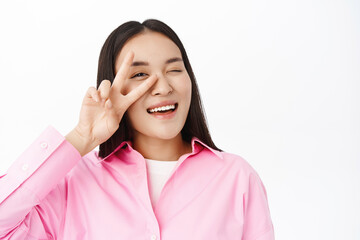 Close up of asian girl shows peace sign near face, winks at you and smiles, stands in pink shirt over white background