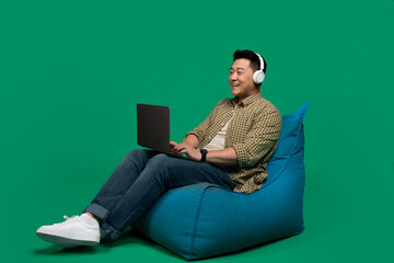Asian man using laptop in headphones, working or having online meeting, sitting in beanbag chair over green background