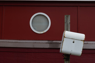 Selective focus outdoor exterior at white post box in front of red boat house wall and circle window.