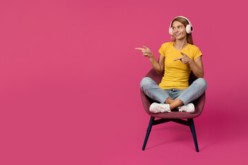 Fototapeta na wymiar Positive middle aged european female sit in armchair in lotus position, listen music in wireless headphones