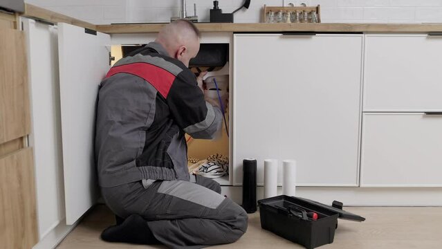 Repairman in overalls installs or changes drinking water filters under the sink in the kitchen. Service concept household filtration system. Timely replacement of water cartridges is healthy life