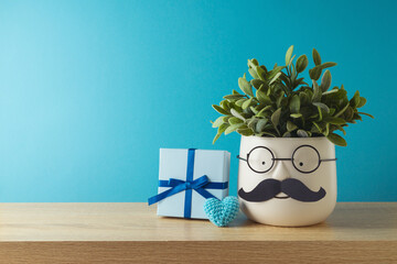 Happy Father's day concept with cute funny plant with mustache, eyeglasses and gift box on wooden table over blue background