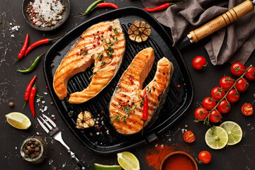 Two grilled salmon steaks in cast-iron pan with vegetables and spices on dark concrete background. Top view, close up