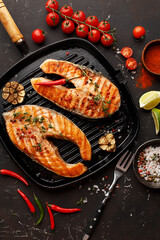 Two grilled salmon steaks in cast-iron pan with vegetables and spices on dark concrete background. Top view, close up