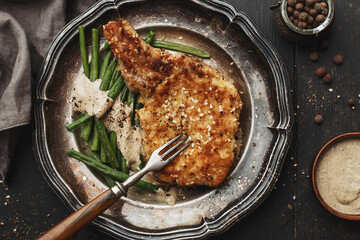 Breaded pork chop with green beans and pepper sauce in vintage plate on dark wooden background. Top view, close up