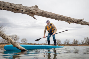 senior male paddling a stand up paddleboard on a lake in early spring, frog perspective from an action camera at water level