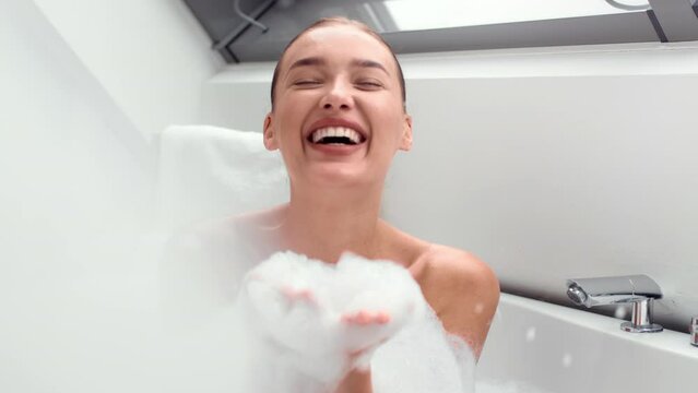 Close up portrait of young happy woman having hot bath at home, blowing soap foam to camera and laughing, slow motion