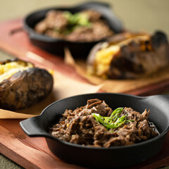 Stewed meat with baked potatoes. Meat in portion pan on wooden board. Lunch at restaurant. Homemade meals. Side view. Second portion of dish is in background. Blurred background. 