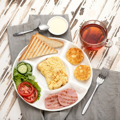 Breakfast plate. Omelet, cheesecakes, fried sausage, vegetables, herbs, toasted bread. Breakfast, tea, yogurt and cutlery on wooden table. Homemade breakfast. View from above. Wooden background. 