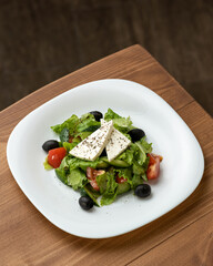 Salad with vegetables and feta cheese. Greek salad with olives, feta, lettuce, cucumber and tomatoe. Healthy, low calorie food. Fitness menu. Dish on white plate on wooden table. Top view. Copy space.