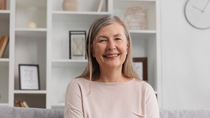 Happy senior woman sitting on sofa in nice apartment. A grayhaired grandmother in her 50s or 60s is resting on the couch at home, smiling.
