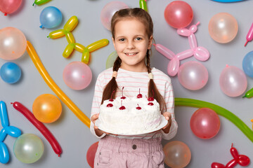 Fototapeta na wymiar Smiling cheerful birthday little girl with pigtails standing with party cake in hands celebrating holiday against gray wall decorated with colorful balloons.