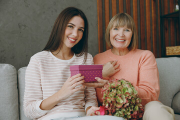 Two happy adult women mature mom young kid wear casual clothes present box with gift ribbon bow sit on gray sofa couch stay at home flat rest relax spend free spare time in living room Family concept