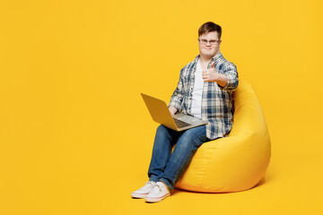 Full body young IT man with down syndrome wear glasses casual clothes sit in bag chair work hold use laptop pc computer show like isolated on plain yellow background Genetic disease world day concept