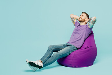 Full body side view happy young man he wears purple t-shirt sit in bag chair hold hands behind neck isolated on plain pastel light blue cyan background studio portrait Tattoo translates life is fight