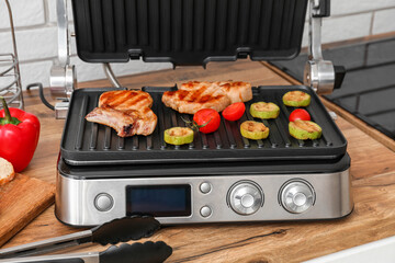 Modern electric grill with tasty steaks and vegetables on table near light brick wall