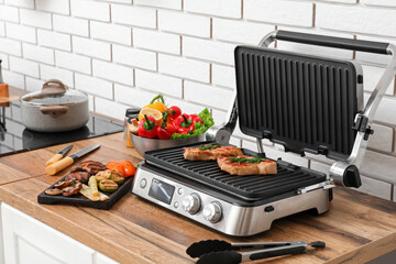 Modern electric grill with tasty steaks and vegetables on table near light brick wall