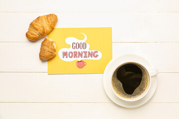 Cup of coffee, tasty croissants and card with text GOOD MORNING on light wooden background