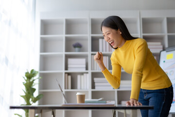 Successful asian business woman happy with laptop computer at home. Freelancer Asian woman yelling yes, looking at the laptop screen, celebrating victory, goal achievement raised arms up.