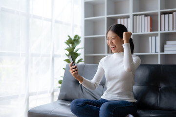 Young Asian female reading good news on phone. Surprised asian woman celebrating victory on smartphone on sofa at home.