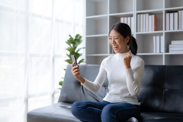 Young Asian female reading good news on phone. Surprised asian woman celebrating victory on smartphone on sofa at home.