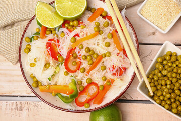 Composition with bowl of tasty rice noodles, vegetables, sesame seeds and lime on light wooden table