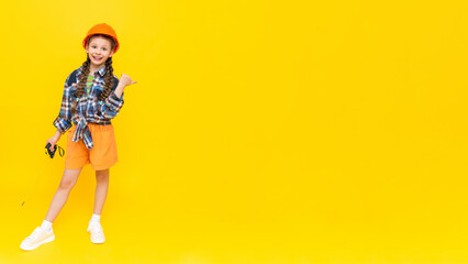 A child in a construction helmet points to your advertisement. A happy little teenage girl in full height holds a construction. A place for advertising. Yellow isolated background. Space for copying.