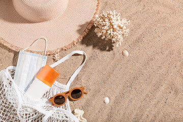 Fototapeta na wymiar Bag with bottle of sunscreen cream, medical mask, sunglasses, hat and coral on sand. Melanoma concept