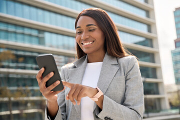 Young smiling successful African American business woman, beautiful middle age businesswoman using smartphone, cellphone application walking at city urban street in front of business center outdoors.