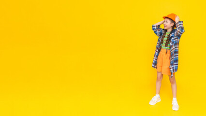 A child in a construction helmet points to your advertisement. Happy little teen girl in full height. A place to advertise. Yellow isolated background. Copy space. Banner.
