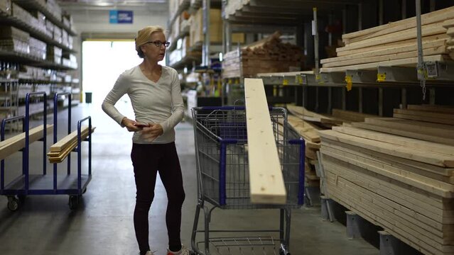 Attractive mature elderly blonde woman using smart phone in lumber section of hardware store. Concept of older person shopping for home improvement experience